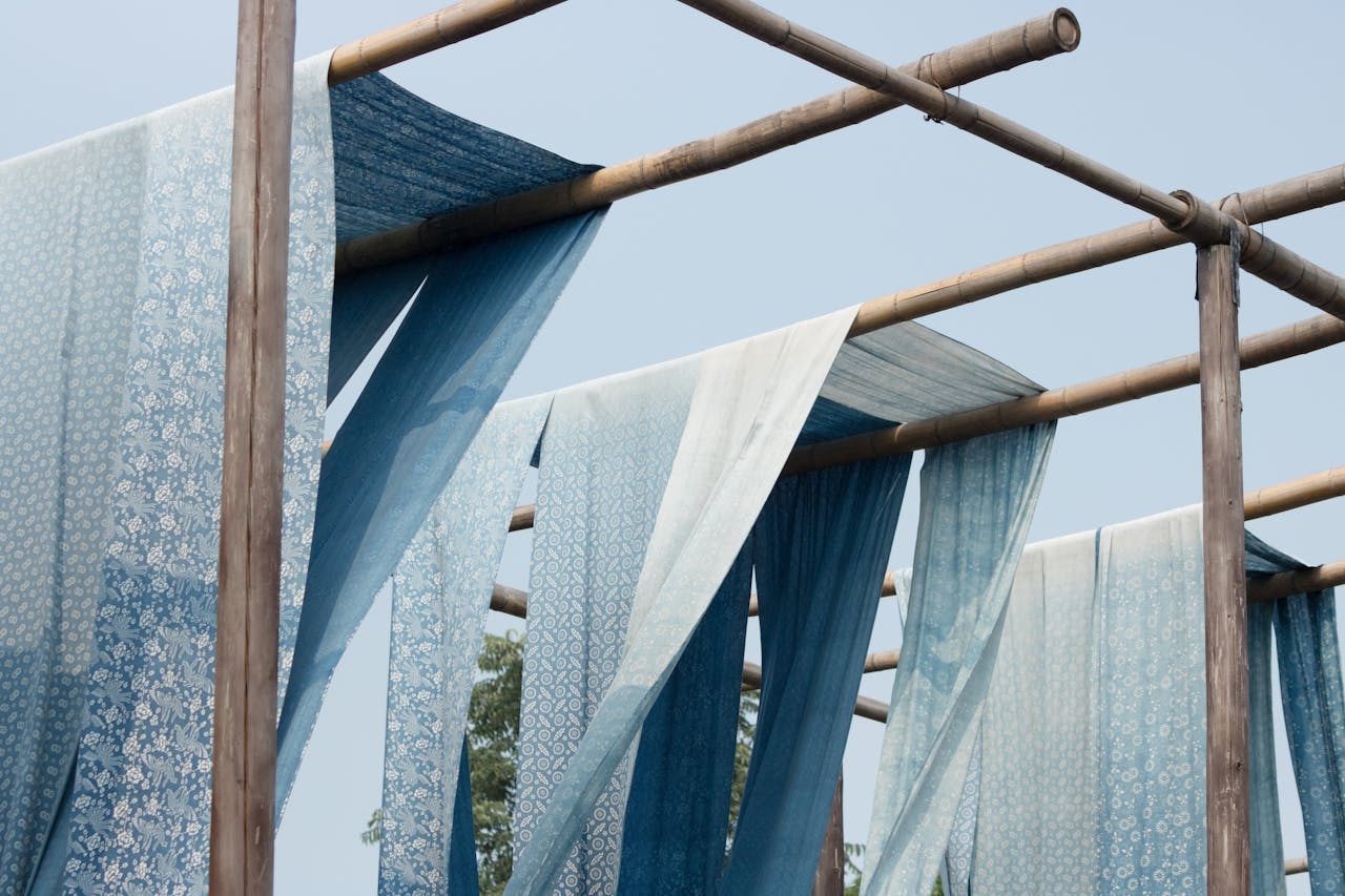 Blue patterned textiles drying under a clear sky on wooden bamboo frames outdoors.