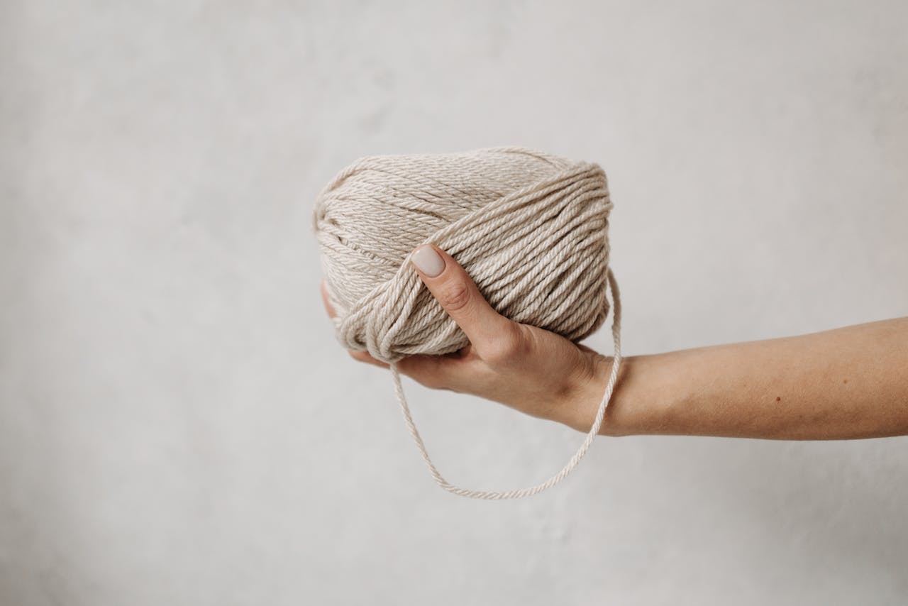 A close-up of a hand gracefully holding a soft beige yarn ball against a neutral background.